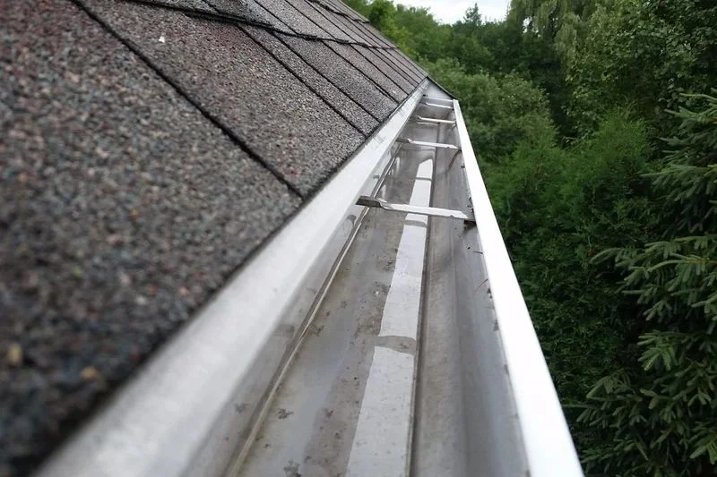 Clean rain gutter running along a shingled roof beside trees